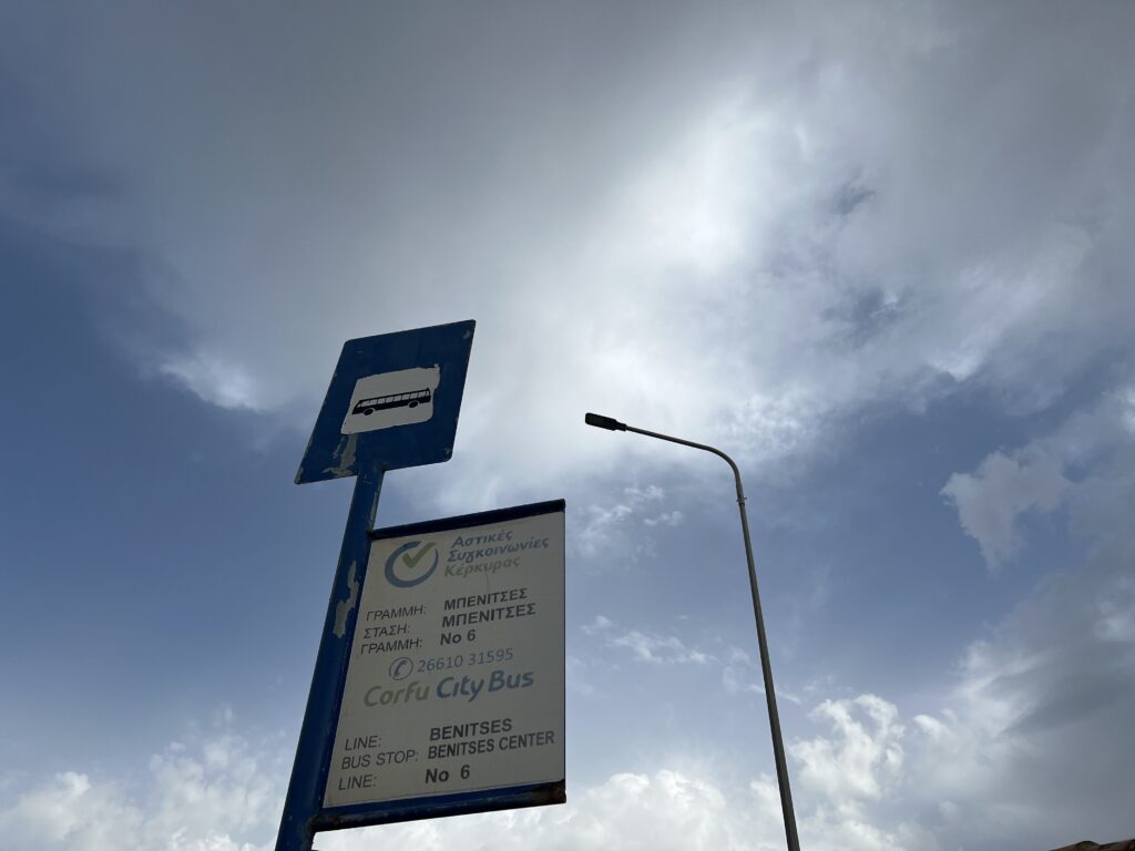 Looking up at a Corfu city bus bus stop sign with blue skies and clouds in the background. The seperate sign above with a bus on it helps finding bus stops and getting around Corfu super easy