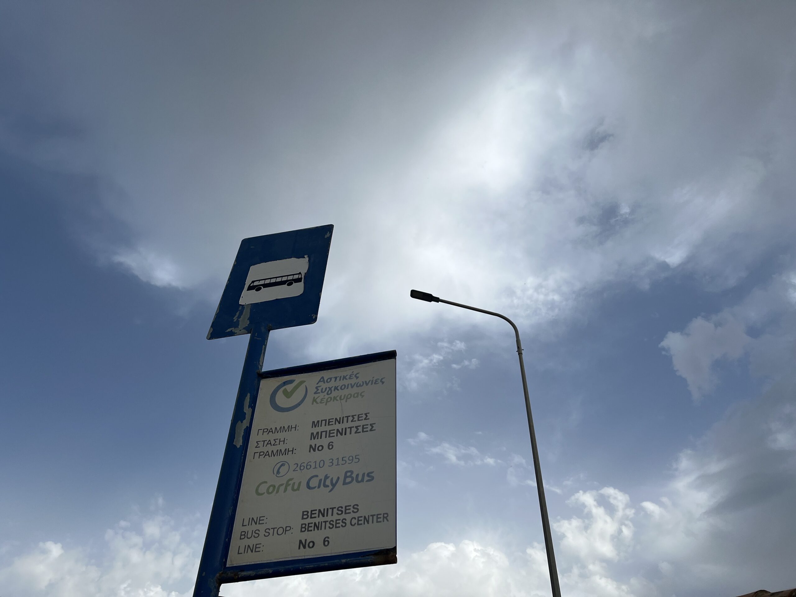 Looking up at a Corfu city bus bus stop sign with blue skies and clouds in the background. The seperate sign above with a bus on it helps finding bus stops and getting around Corfu super easy