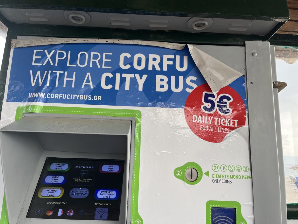 a city bus ticket machine in Corfu. The blue and white banner says "explore Corfu with a city bus" the machine is also advertising a 5 euro day ticket above the screen