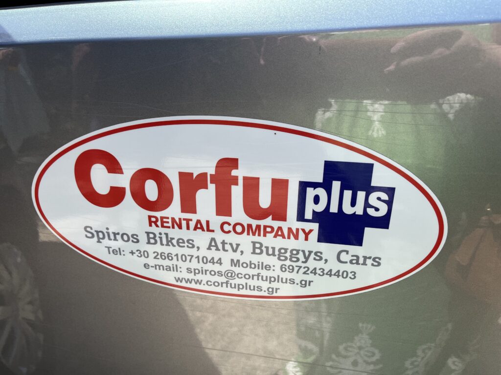A silver car with an oval white sticker on it advertising Corfu Plus rental company