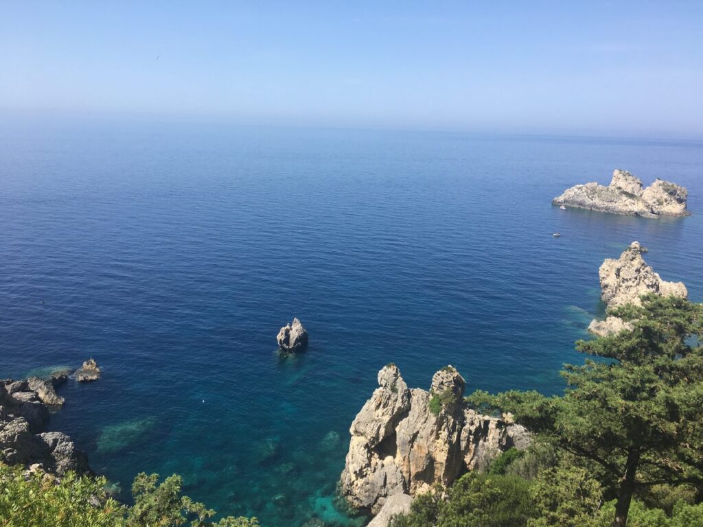 Scenic coastal view of Corfu Island featuring clear blue waters, rugged rock formations rising from the sea, and lush greenery in the foreground under a bright, cloudless sky. Places like this on the island are full of bugs at night!