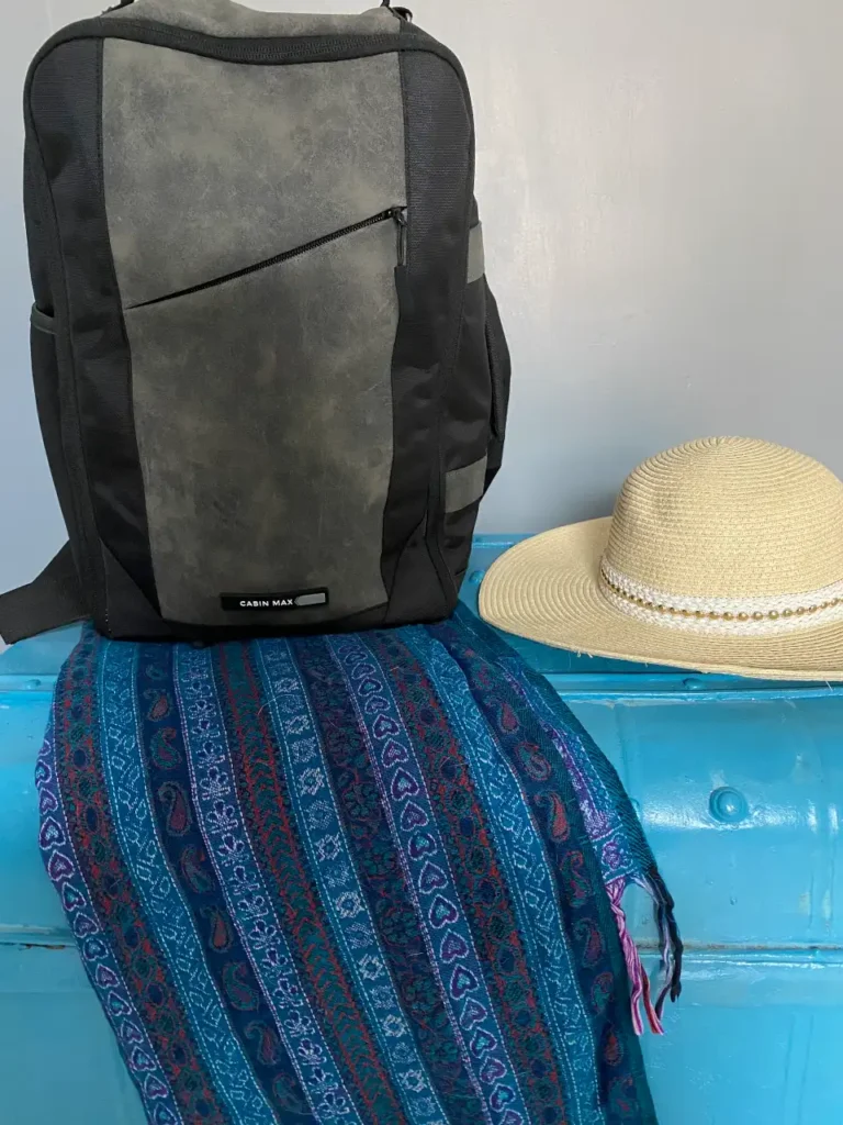 A compact black and grey travel backpack labeled "Cabin Max" rests on a bright blue suitcase, accompanied by a woven blue and purple patterned scarf and a beige straw sun hat with a pearl band—perfect essentials for a summer trip to Corfu