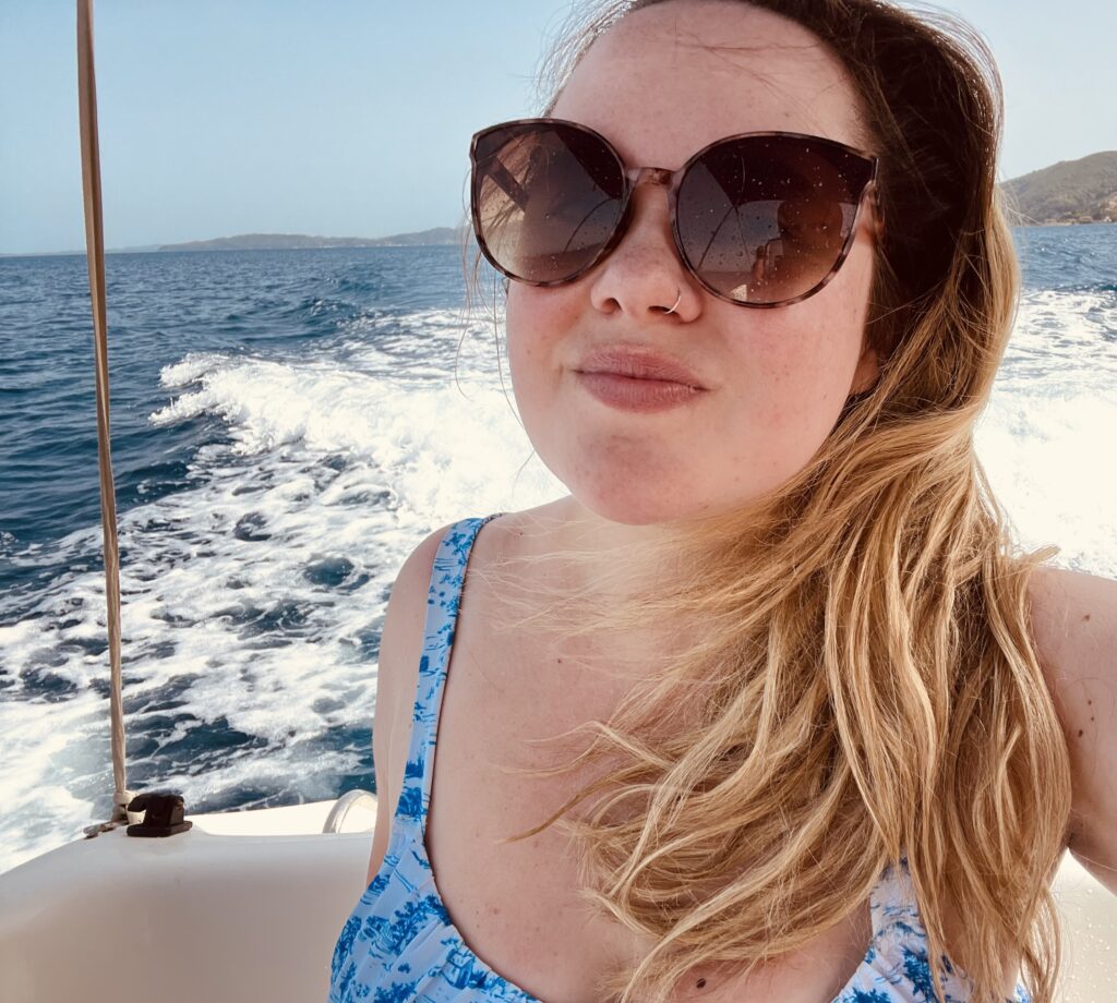 Woman wearing sunglasses and a blue swimsuit smiling while on a boat in Corfu, with waves trailing behind. This image is part of a blog post sharing a personal experience of hiring a boat and exploring the coast of Corfu