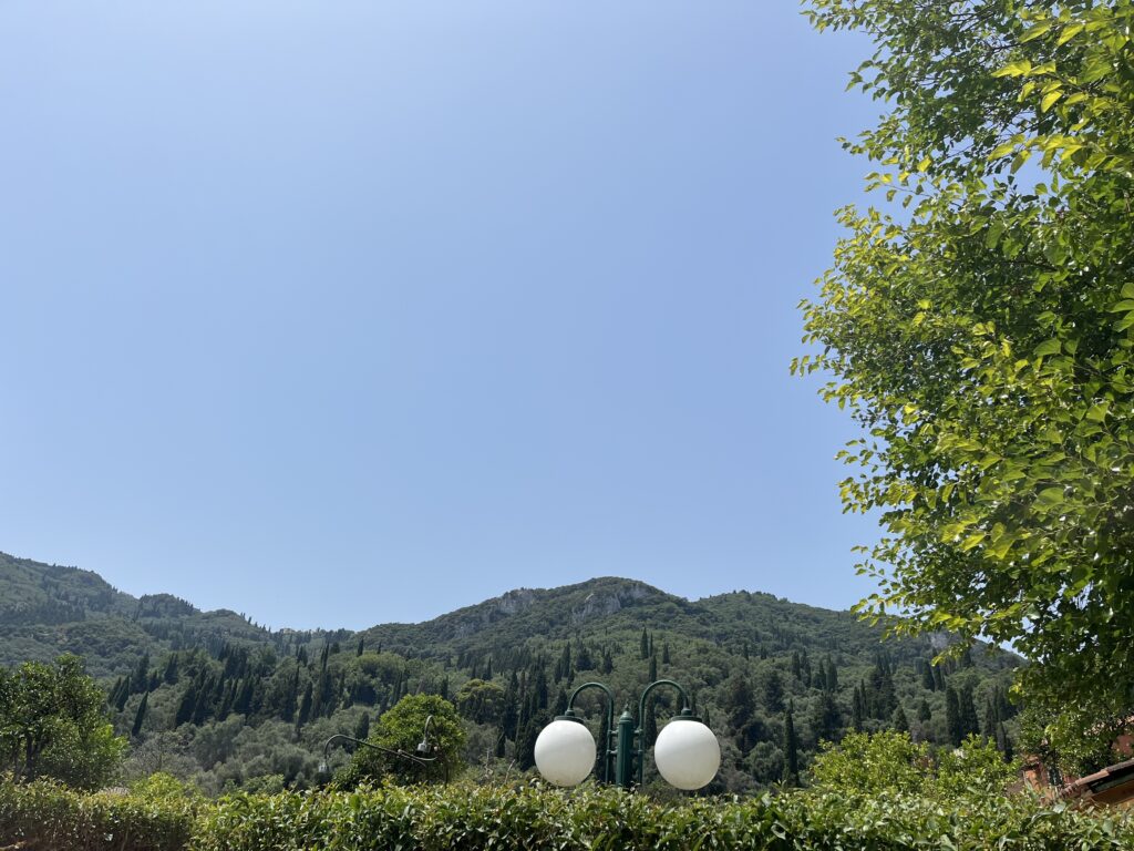 View of green hills and cypress trees under a clear blue sky in Benitses, Corfu. A vintage-style streetlamp and lush greenery are in the foreground. This peaceful village is one of the best places to stay in Corfu without a car.