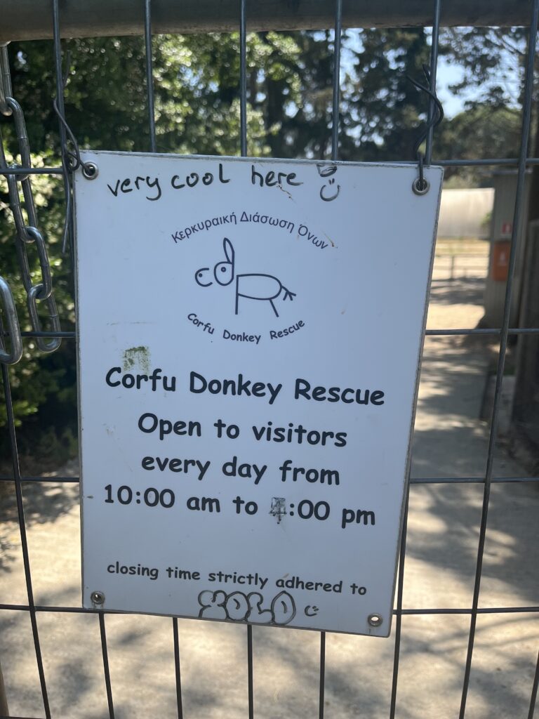 A sign at the entrance of Corfu Donkey Rescue near Doukades displays the opening hours, stating it’s open to visitors every day from 10:00 am to 4:00 pm, with “very cool here” handwritten at the top.